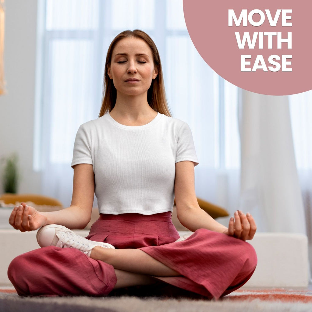 A lifestyle photo of a woman sitting cross-legged in a yoga pose on a grey rug, eyes closed with a peaceful expression. She wears a white cropped t-shirt and pink trousers. A pink circle in the top-right corner features the white text 'MOVE WITH EASE'.