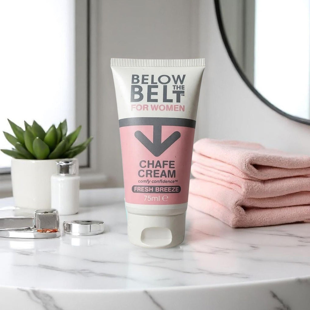 A 75ml tube of Below the Belt for Women 'Fresh Breeze' Chafe Cream standing upright on a white marble countertop. The pink and white tube is surrounded by soft lifestyle elements, including a stack of pink towels.