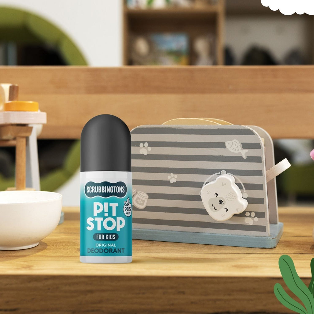 A lifestyle shot of Scrubbingtons Pit Stop Original Deodorant for Kids. The 75ml roll-on bottle is displayed on a wooden surface next to a grey-and-white striped toy carry case in a brightly lit playroom or classroom setting