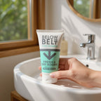 A hand holding a tube of Below the Belt for Women: Fresh & Dry Boobs anti-chafing cream.  The product is positioned over a modern white bathroom sink in a bright, sunlit room