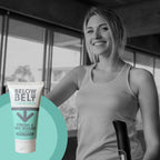 A smiling woman in a gym setting using an elliptical machine. In the foreground, a vibrant, full colour tube of Below the Belt for Women: Fresh & Dry Boobs