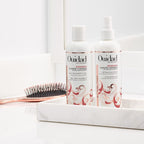 Two bottles of Ouidad hair care products on a white surface with a brush in the background.
