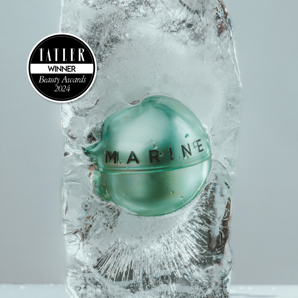 A close-up of a round, translucent aqua-green cosmetic container sealed inside a large block of ice, with the brand name MARINE embossed on the front. A black circular 'TALLER WINNER Beauty Awards 2024' badge appears in the upper left.