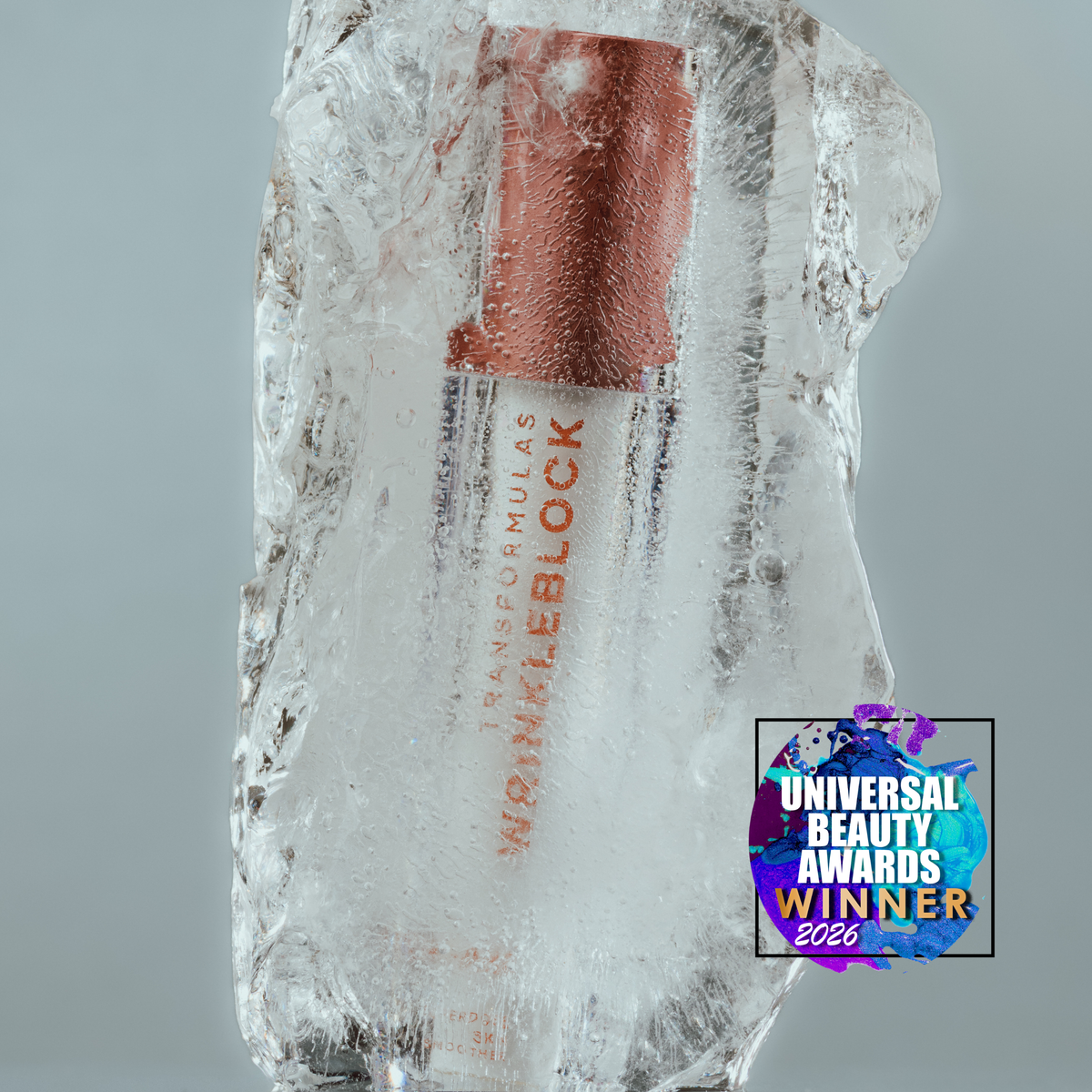 A lip gloss tube wrapped in clear ice, with a copper-toned cap and red text reading 'TRANSCUTANEOUS WRINKLEBLOCK' and 'LIP GLOSS'. A 'Universal Beauty Awards Winner 2026' badge is shown in the lower right corner against a neutral gray background.